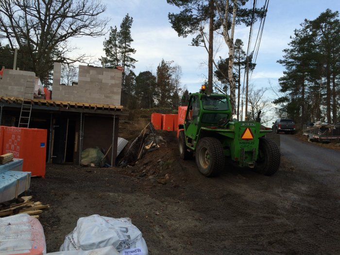 Byggarbetsplats med teleskoplastare, blockpallar nära en halvfärdig byggnad och byggmaterial på marken.