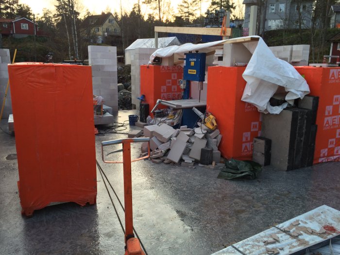 Byggplats med staplade byggblock och byggmaterial, skyddade av orange presenningar.