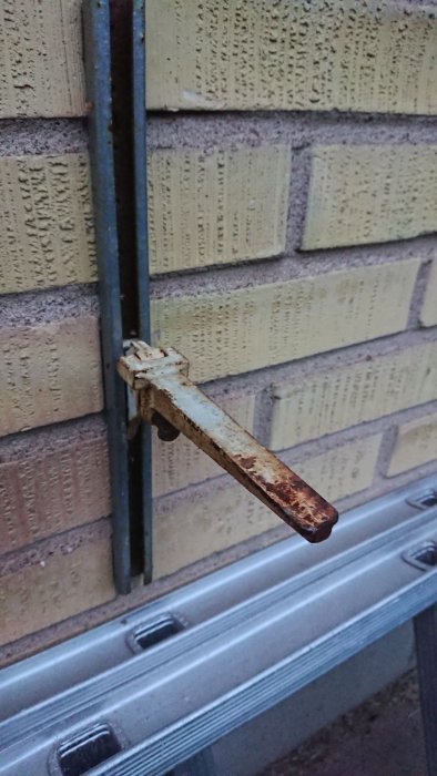Rostig metallhållare för stege monterad på en tegelvägg med en del av en stege synlig.