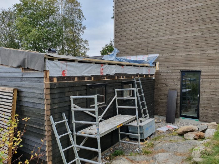 Byggprojekt med nylagd panel och vindskydd vid taket, arbete på balkongen syns, olik färg på panelen.