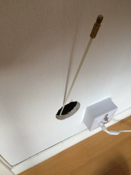 Borrat hål i vägg med synlig rotdosa och kabel genom mellanbjälklaget.