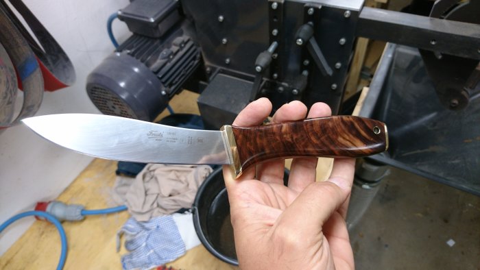 Nyputsad jaktkniv med glänsande blad och polerat trähandtag hålls framför en verkstadsutrustning.