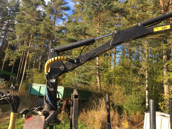 Kranarm på skogsmaskin framför träd och en grönskande skogsbakgrund.