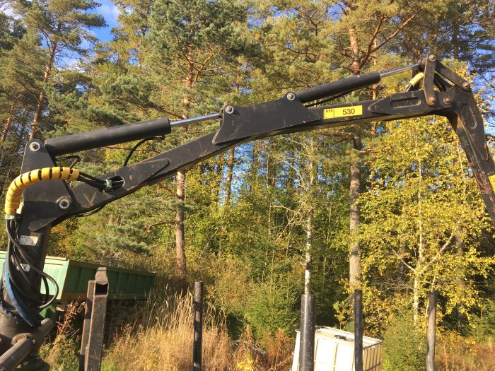 Grävmaskinsarm i skogen med synlig modellbeteckning 530, i bakgrunden lastbil och träd.