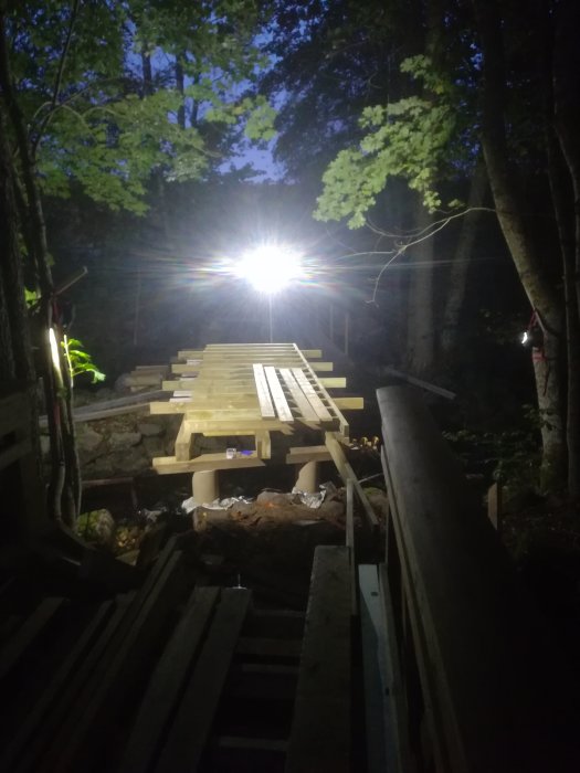 Batteridriven strålkastare lyser upp en nybyggd träbro i nattmörker omgiven av skog.