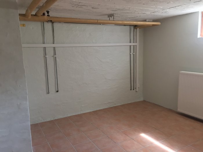 Källarrum med vita väggar, tegelröda klinkers och synliga rör längs taket och väggen innan isolering med glasull.
