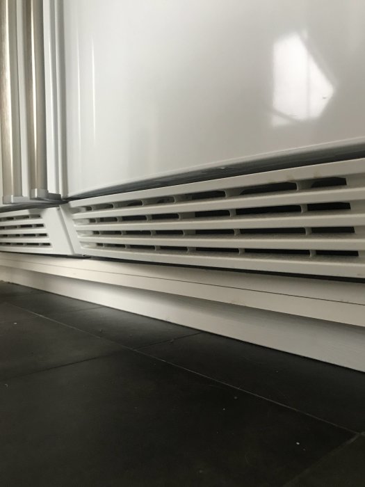 Vit sockel under skåp med ventilationsgaller mot ett mörkt golv.