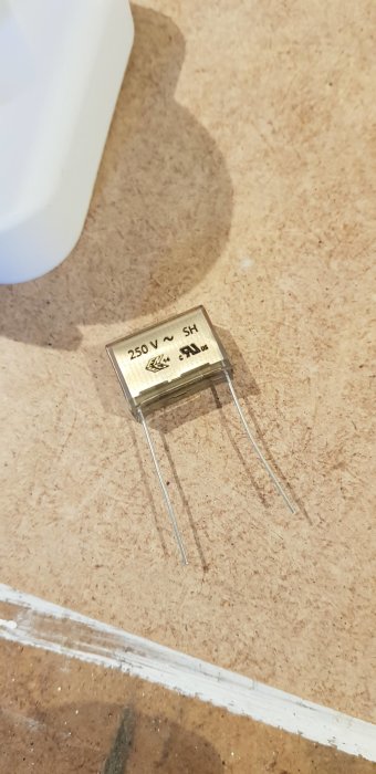 Kondensator med beteckningen 250V SH ligger på ett brunt bordsunderlag.