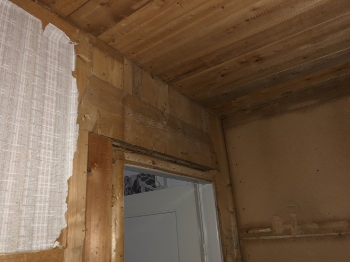 Veggen innendørs med synlig treverk og delvis avrevet tapet rundt en dørkarm, sannsynligvis en bærende vegg som skal rives.