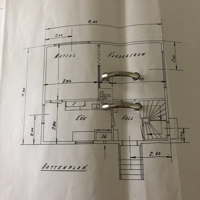 Planløsning av et hus fra 1950 med markerte mål for forskjellige rom, inkludert matsal, kjøkken og vindfang. To metallhåndtak vises.