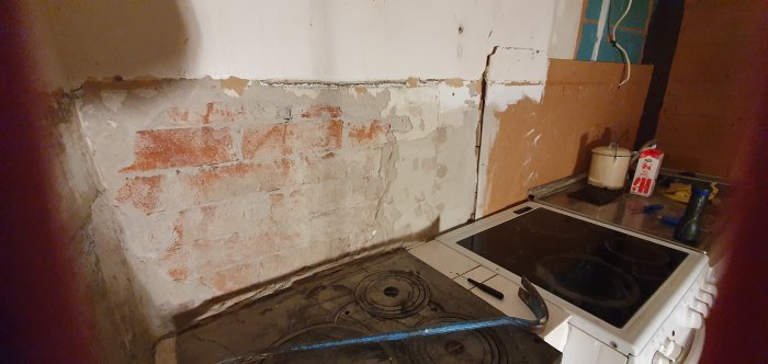 Ett kök under renovering med avlägsnat kakel och ojämn väggyta av tegel och puts ovanför en äldre spis.