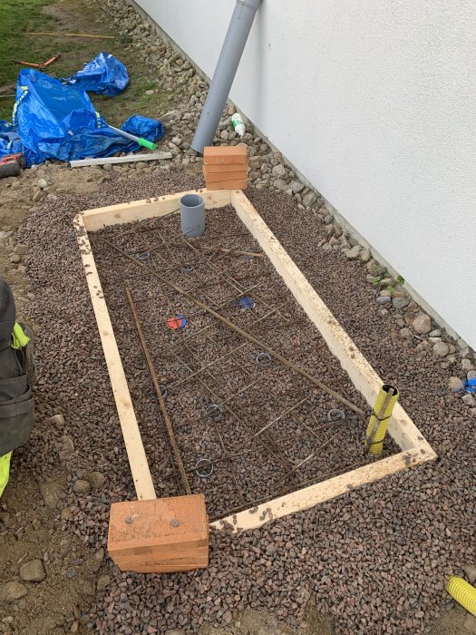 Nybyggd betongplatta med armeringsjärn, träformar och grus runtomkring, vid en husvägg.