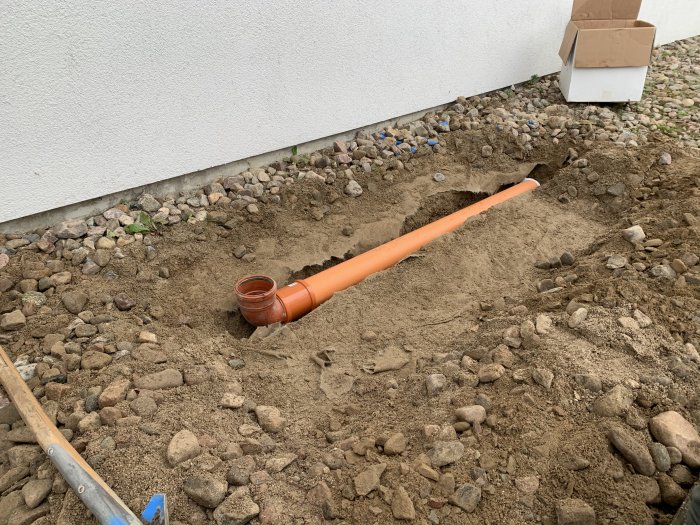 Nyinstallerad orange avloppsrör i grävd ränna bredvid husvägg med jord och stenar.