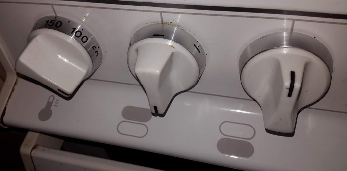 Äldre spisens vitklädda vred för temperaturkontroll och undervärme, visar slitage och smuts.