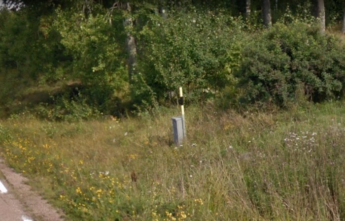Suddig bild av en grå låda med gul/svart pinne vid en tomtgräns i ett grönt område.