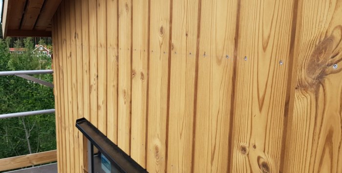 Nyinstallerad Thermowood fasad med gyllengul ton och synliga skruvar mot en balkong och grönska i bakgrunden.