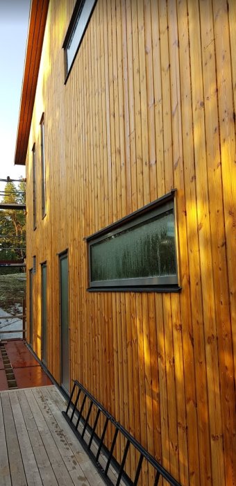 Solnedgången ger en varm brun-röd ton till den gula Thermowood-fasaden på ett hus med fönster och reling.