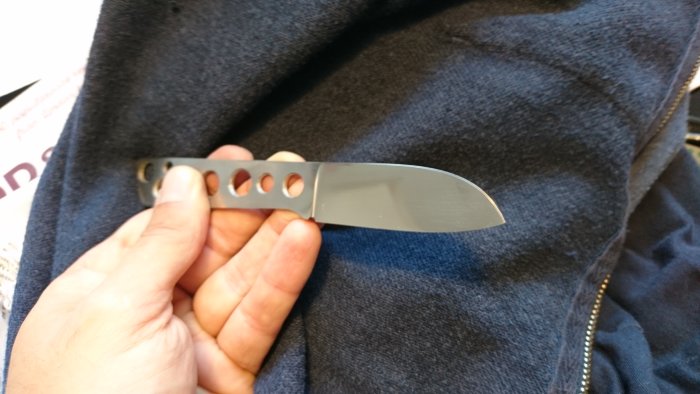 Hand håller ett grovpolerat knivblad i RWL 34 stål med modifierad "fårfot"-design och hål.