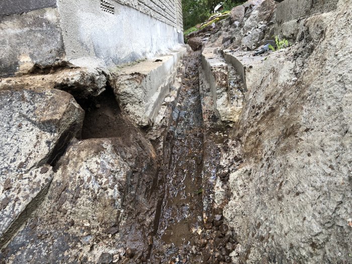 Ett dräneringsschakt skuret intill husgrund på berg med synliga skador och porös betong, omgivet av grus och vattenrännor.