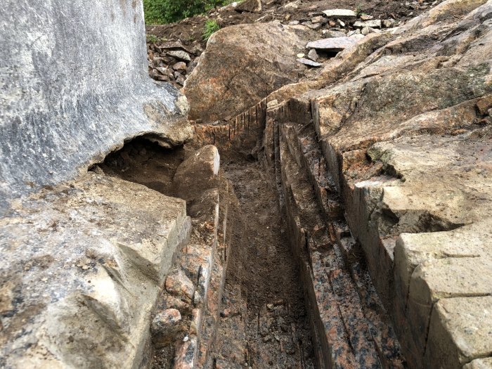 Dräneringsschakt med synliga sprickor och skador i grundens betong bredvid berggrund, visande tecken på erosion och vattenskador.