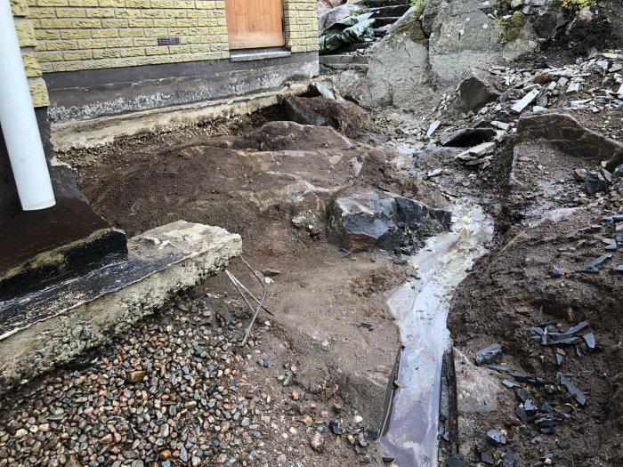 Dräneringsschakt vid husgrund med synligt berg, grus och delar av betonggjutning i olika stadier av reparation.