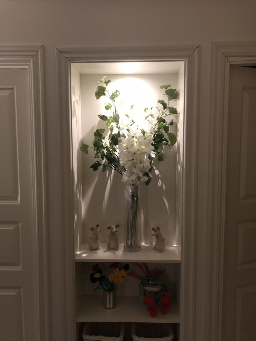 Fönsternisch med belyst vit blomarrangemang i vas samt figurer och en metallburk med blommor.