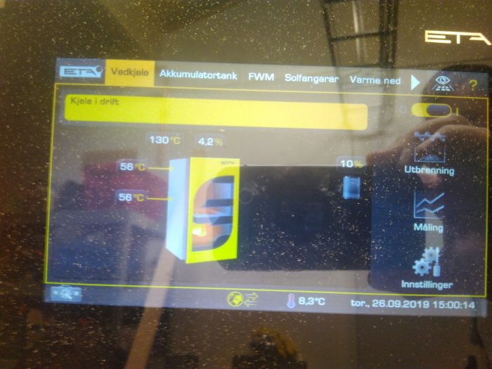 Kontrollpanel för en vedpanna som visar driftstatus och temperaturer, med smutsigt glas framför.