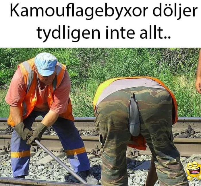 Arbetare i hi-vis kläder och kamouflagebyxor böjer sig ner med synlig reva på byxorna medan en annan arbetar på järnvägsspår.