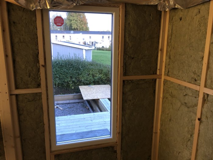 Inre vägg med isolering i ett pågående byggprojekt, trästomme och fönster med utsikt till trädgård.