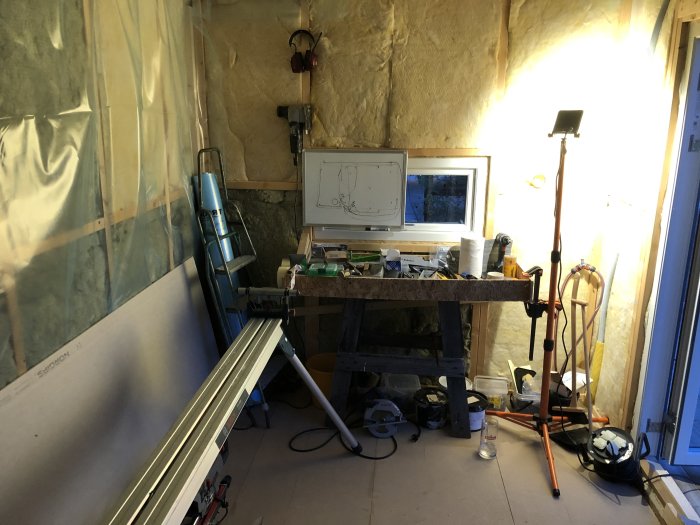 Inre hörn av byggprojekt med gul isolering, verktyg, en arbetsbänk och byggmaterial runtomkring.