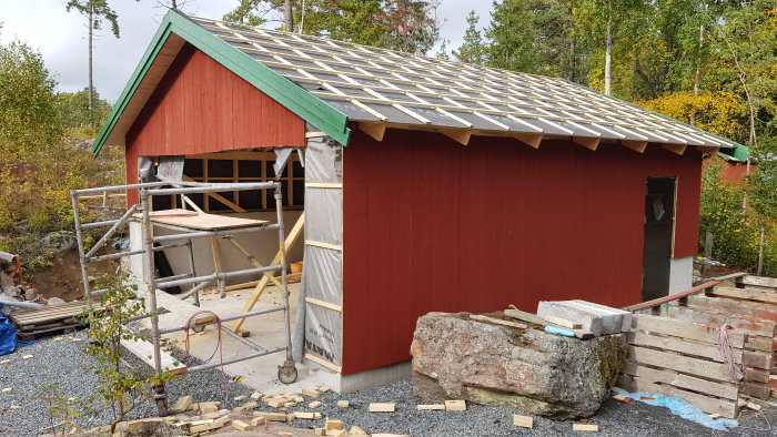 Rödmålat hus under renovering med oskyddad takkonstruktion och underlagsduk.