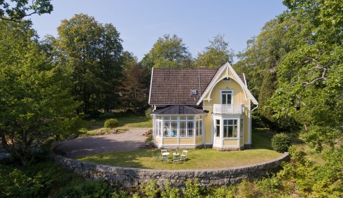 Gult traditionellt hus i Växjö med veranda, omgivet av gröna träd och stenmur.