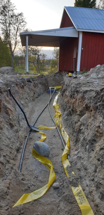 Uppgrävd grävning med varningsband för kabel längs med en slingrande grå kraftkabel, nära en röd byggnad.