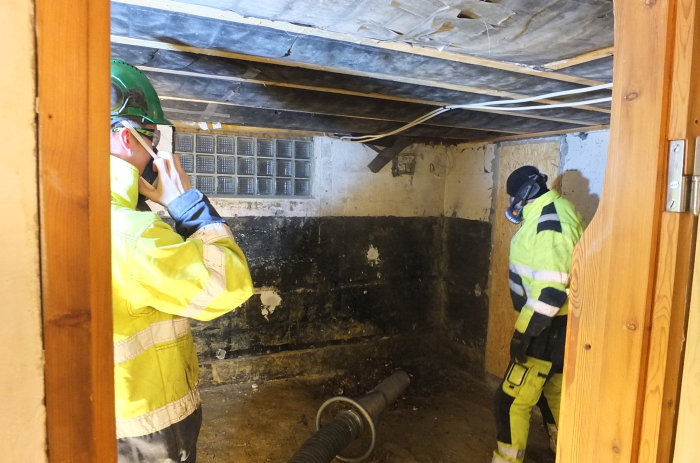 Två arbetare i skyddskläder i ett källarrum med blottade takbalkar och en sugslang på golvet under renovering.