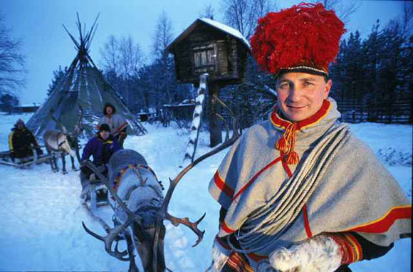 Man i traditionell samisk dräkt framför ren och släde, vinternatur och kåta i bakgrunden.