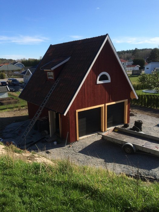 Rött garage med loft och 52 graders taklutning, står öppet med släpvagn framför och byggmaterial synligt.