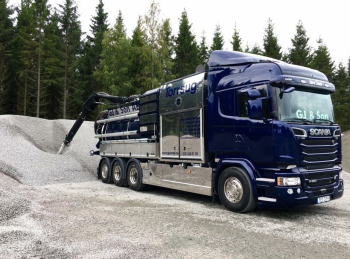 Blå sugbilsenhet med slangen aktiv vid en grushög i skogsmiljö, demonstrerar arbete under fastigheter.