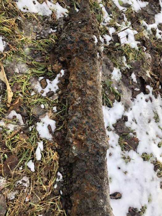 Kraftigt korroderat gjutjärnsrör ligger på marken med snö och gräs som delvis visar igenom.