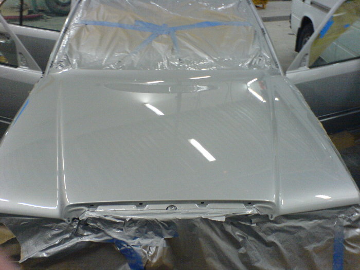 Nylackad vit motorhuv på en klassisk bil i en verkstadslokal, skyddad med plast och tejp.