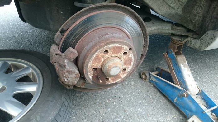 Slitna bromsskivor på bil med rost och vågor på ytan, lyft av en gammal blå domkraft.