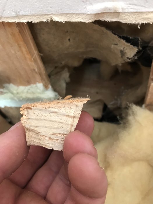 Närbild på en hand som håller en träbit framför en vägg med synlig isolering.