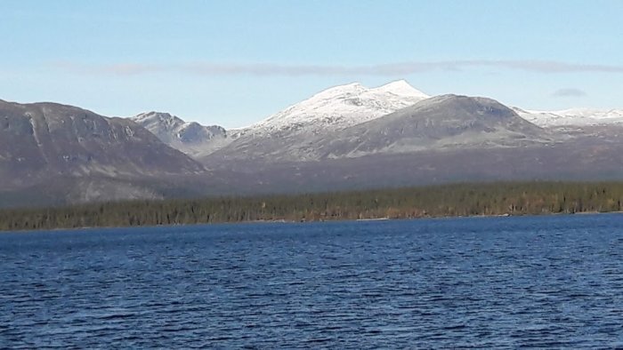 Vy över ett stilla vatten med snöklädda bergstoppar i bakgrunden under en klarblå himmel.