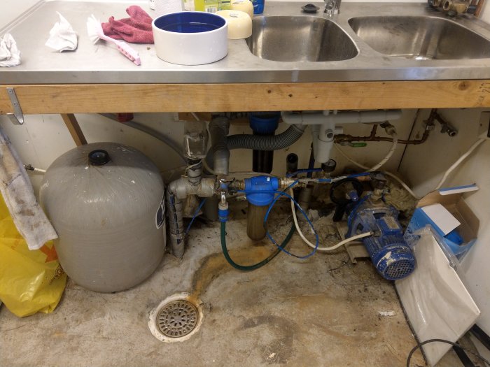 Köksskåp öppet visar vatteninstallation med pump, trycktank och rör under diskho.