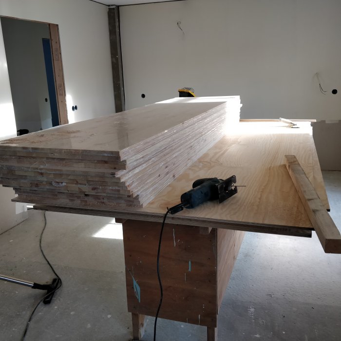 Byggverkstad med stapel av plywood och handöverfräs på ett arbetsbord i halvfärdigt rum.