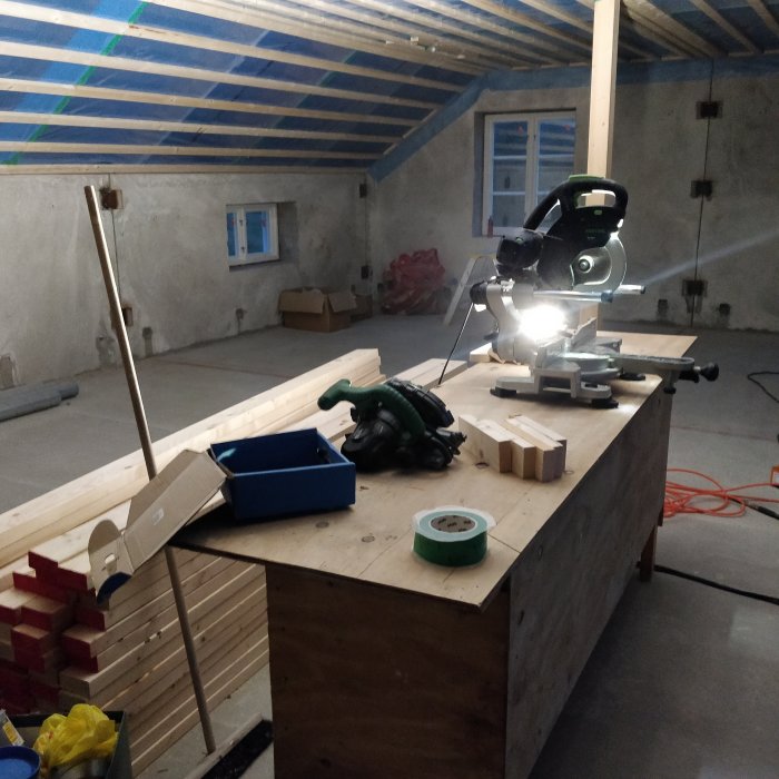 Verktyg och trä på bord i ett byggprojekt inomhus med oisolerade väggar och takstolar.