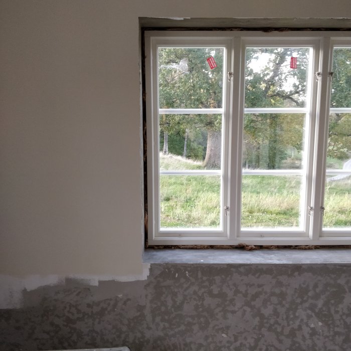 Renoverat rum med nymålade väggar i beige, oputsat område kvar vid fönster med utsikt över natur.