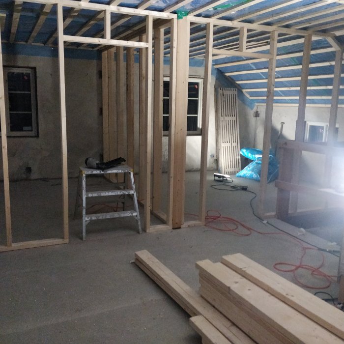 Nybyggda träregelväggar på en övre våning med byggmaterial och stege, ofärdigt byggprojekt under arbete.