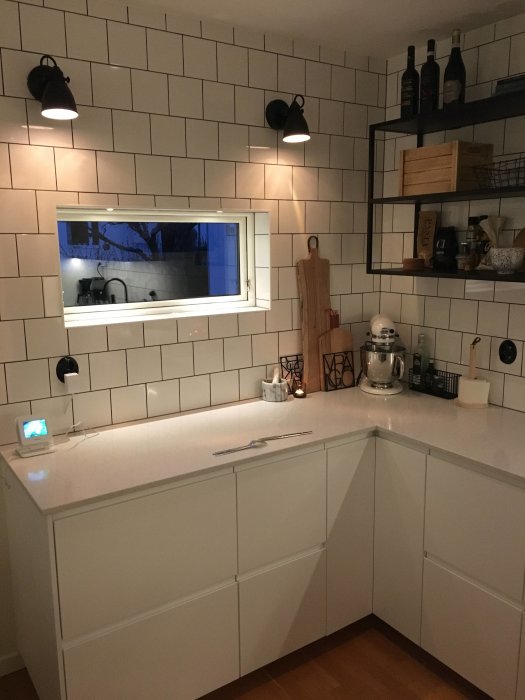 Renoverat kök med vita skåp från Ikea, stenskiva, kakelvägg och svart hyllsystem.