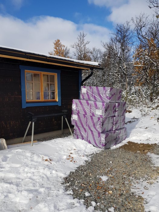 Stapel med isoleringsplattor vid ett snötäckt hus med mörk träfasad och blå fönsterkarmar.