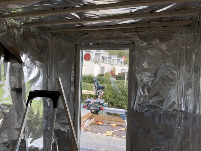 Inuti en bastu under renovering med isoleringsfolie på väggarna och en öppning blickar ut mot en person utanför.